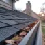 Clogged home gutter filled with dry fall leaves along an asphalt shingle roof, with a brick chimney and suburban house in the background at sunset.