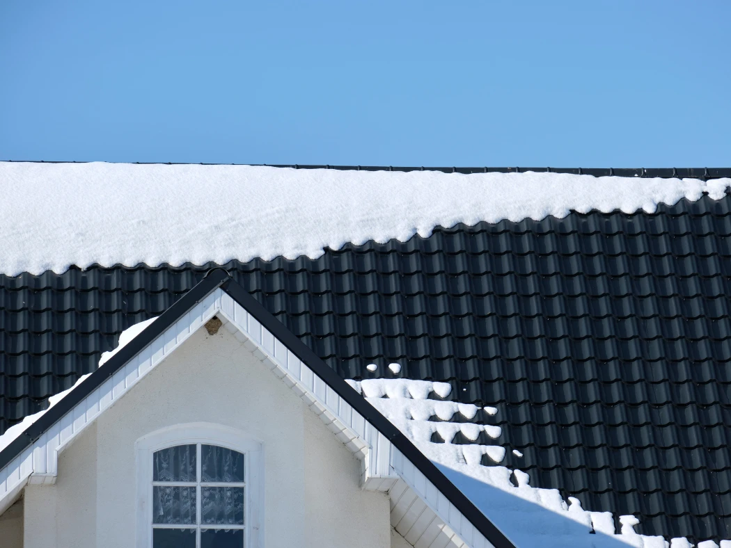 Roof with black shingles on a bright house, top window visible, and snow on the roof—illustrating Holiday Roof Care Homeowners Forget and winter roof maintenance risks.